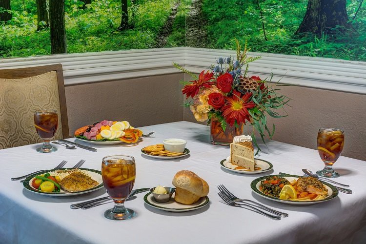 A beautifully set dining table with a variety of dishes, including bread, cake, and iced tea, adorned with a floral centerpiece.