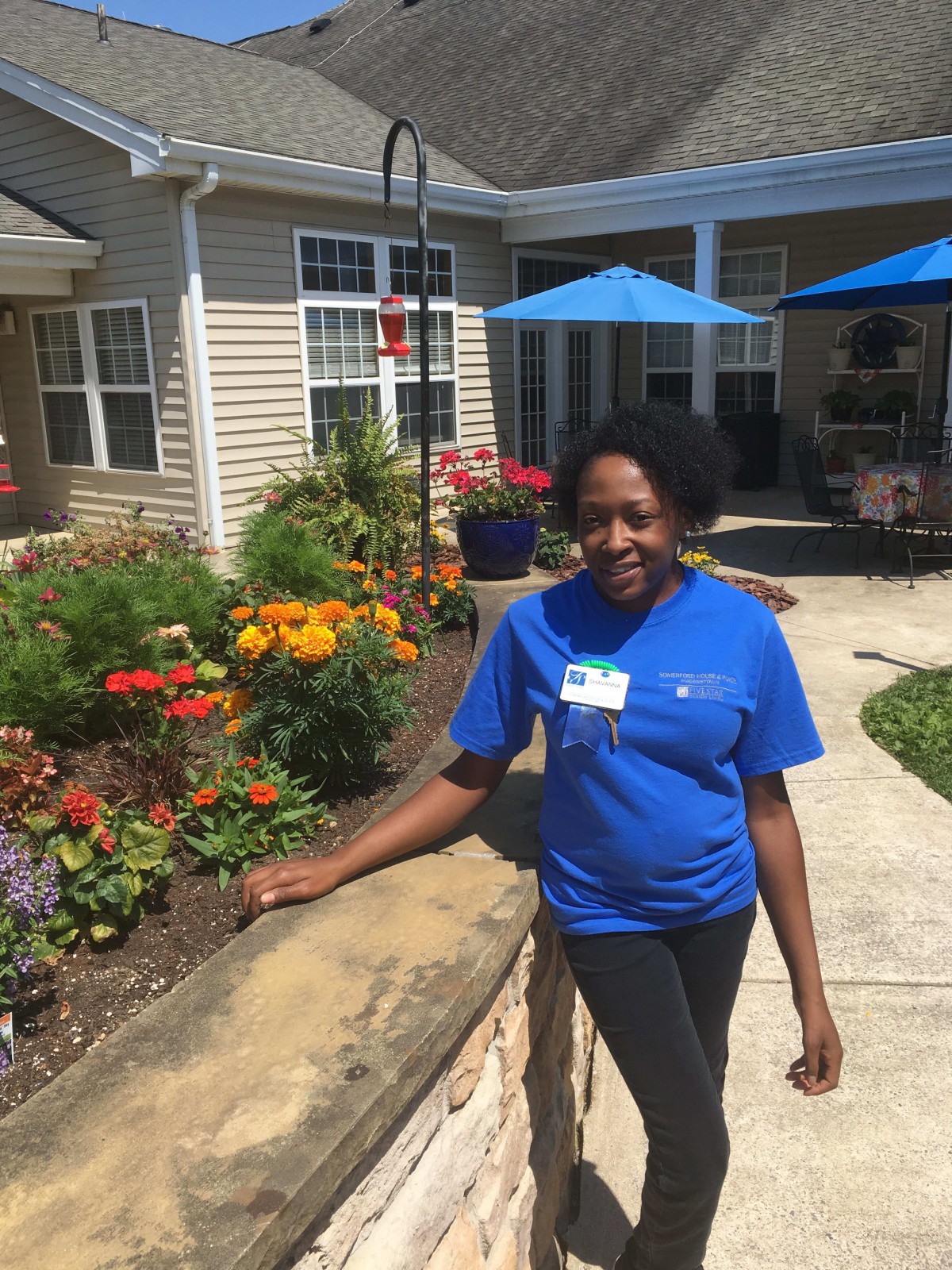 A person standing outside in a garden with colorful flowers and a building in the background.
