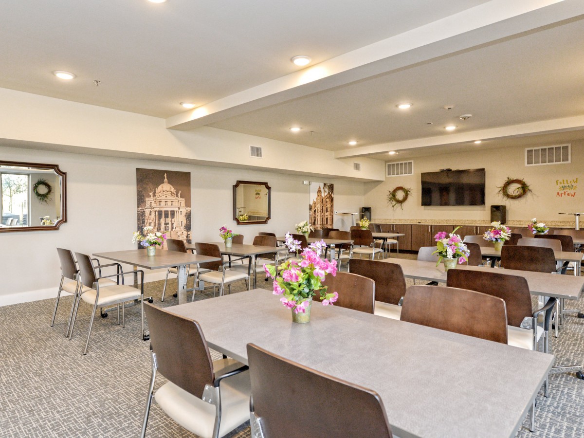 A well-lit community room with tables and chairs arranged neatly, adorned with floral centerpieces.