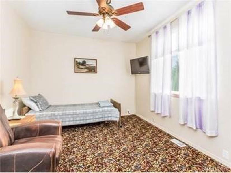 A simple bedroom with a single bed, patterned carpet, ceiling fan, and wall-mounted television.