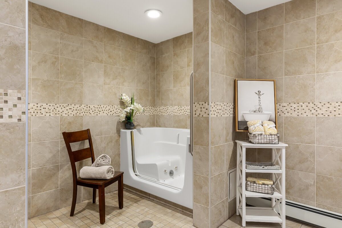 A modern bathroom featuring a walk-in tub, elegant tile walls, and neatly arranged decor with a wooden chair and shelving unit.