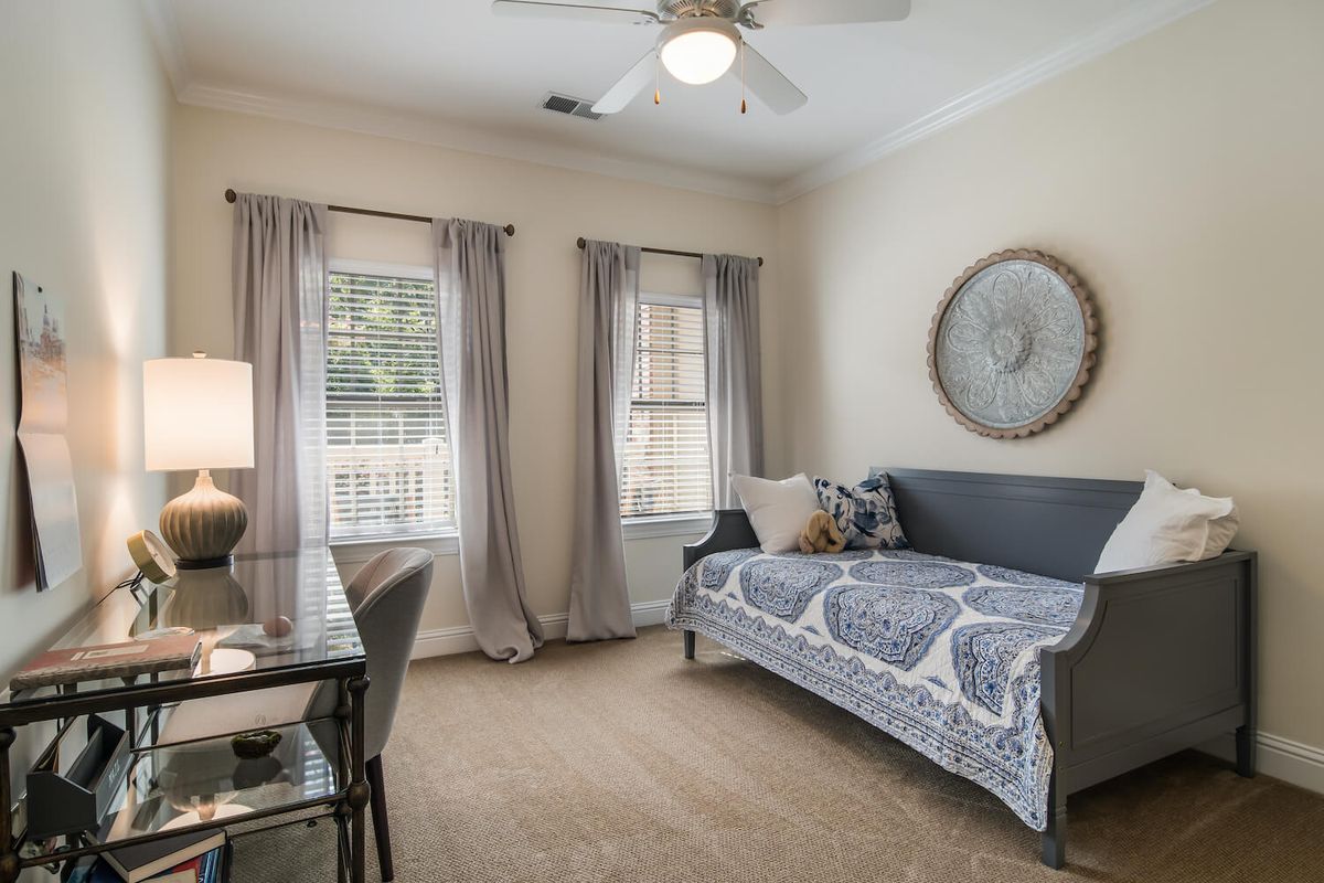 A cozy bedroom with a daybed, desk, and two windows with curtains, featuring a decorative circular wall piece and ceiling fan.