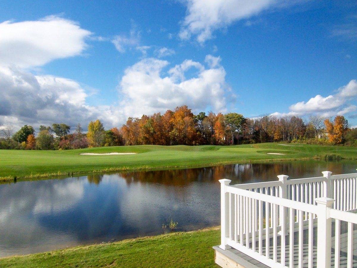 A scenic view of a golf course with a lake, surrounded by autumn trees under a clear blue sky.