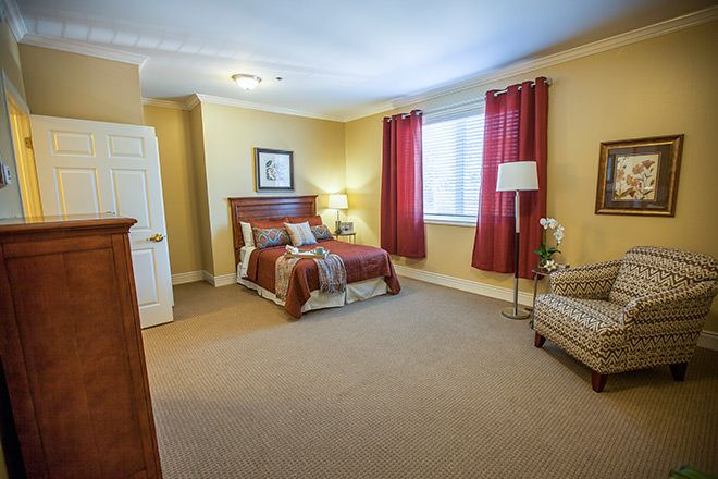 A cozy bedroom with a bed, chair, and table lamp, featuring red curtains and warm lighting.