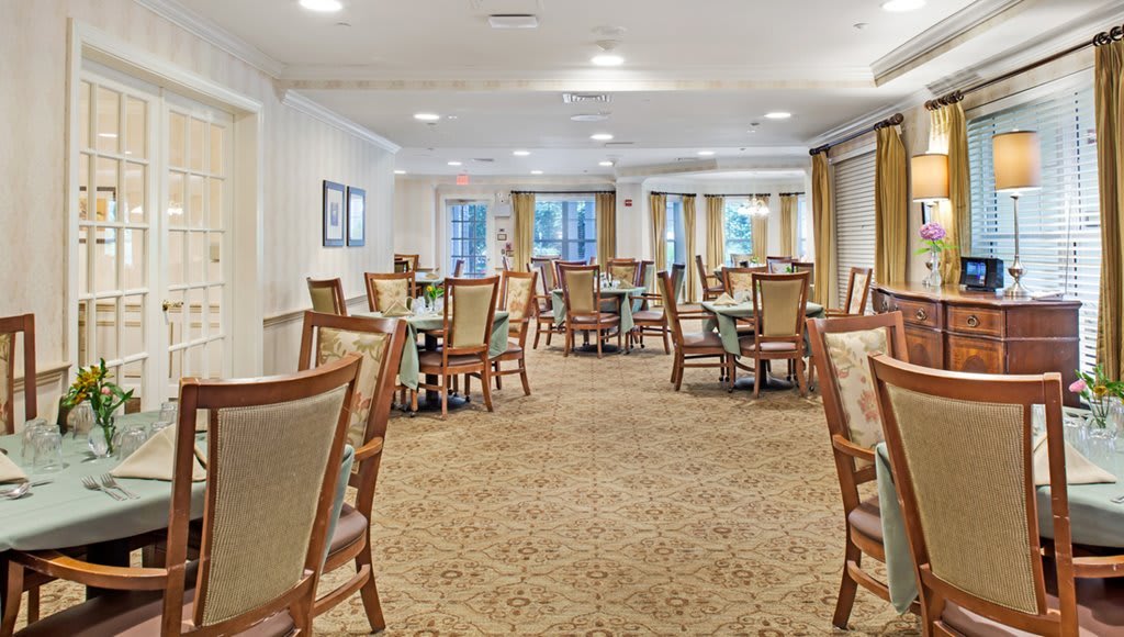 A spacious dining room with neatly arranged tables and chairs, carpeted flooring, and natural light coming through the windows.
