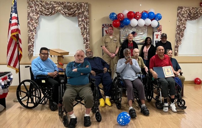 A group of elderly individuals, some in wheelchairs, pose for a photo with uniformed personnel and balloons, suggesting a celebratory event.