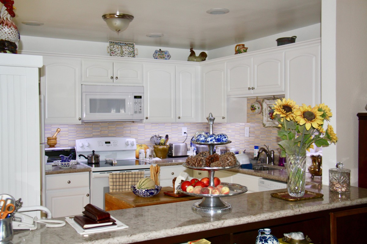 A cozy kitchen with white cabinets and a granite countertop, featuring sunflowers in a vase and a fruit basket.