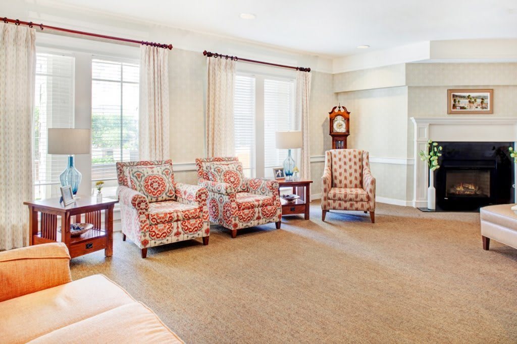 A bright living room featuring floral armchairs, large windows with curtains, and a classic clock.