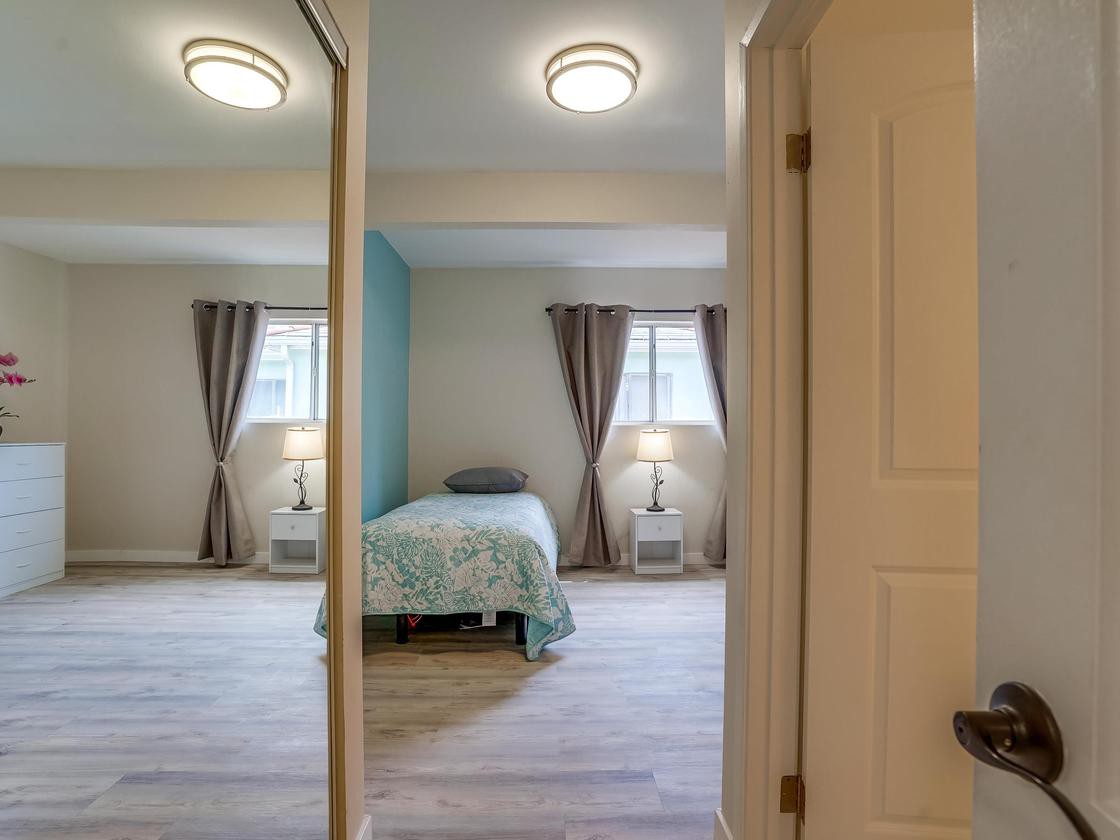 A cozy bedroom with a single bed, mirrored closet doors reflecting the room, nightstands with lamps, and curtains on the windows.