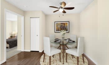 A modern dining room with a round glass table and four white chairs, featuring a ceiling fan and wall art.
