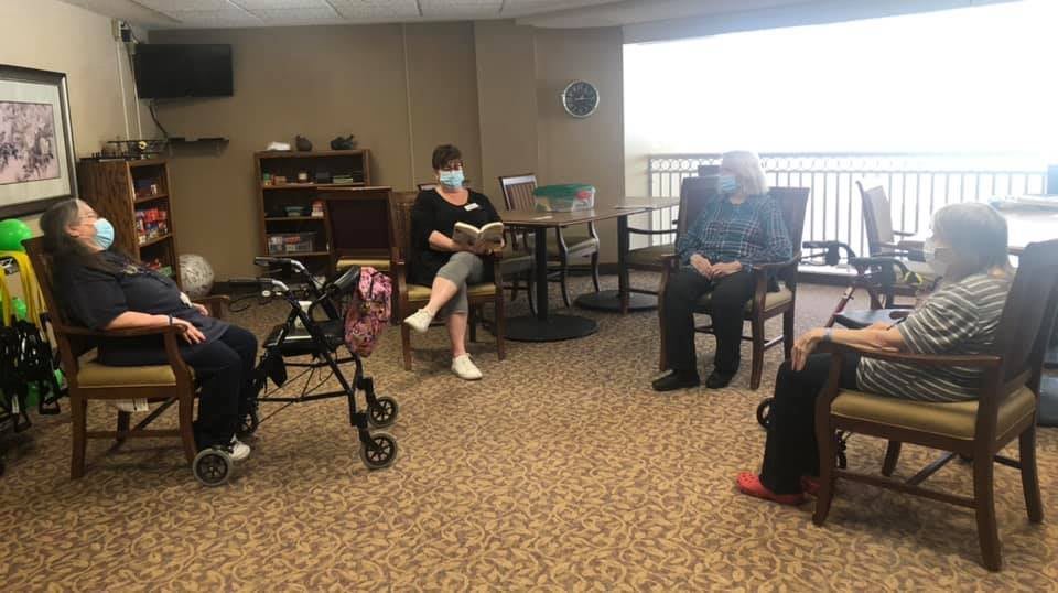 A group of people gathered indoors, with one person reading to the others in a community setting.