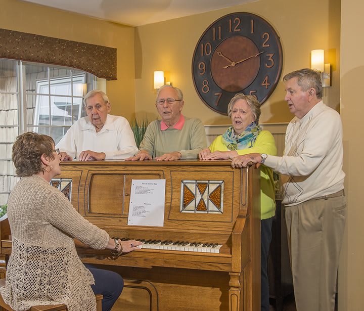 A group of elderly people gathered around a piano, enjoying a singing session indoors.