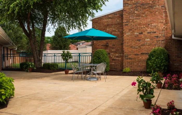 A cozy patio setup with a table, chairs, and a teal umbrella, surrounded by brick walls and greenery.