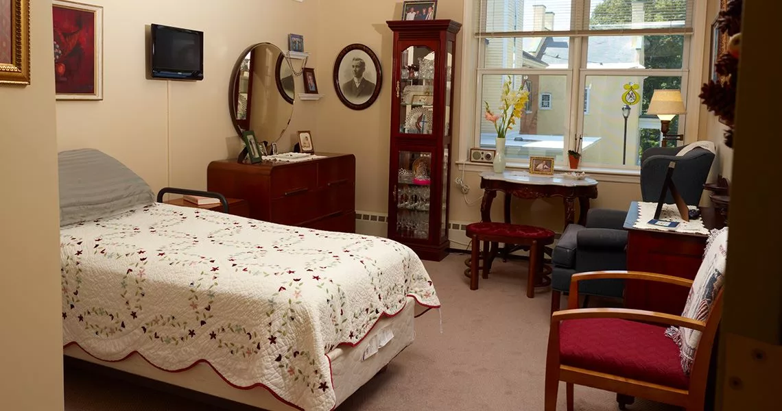 A cozy vintage-style bedroom with classic furniture, including a bed with a lace cover, a dresser with a mirror, and a small table by the window.