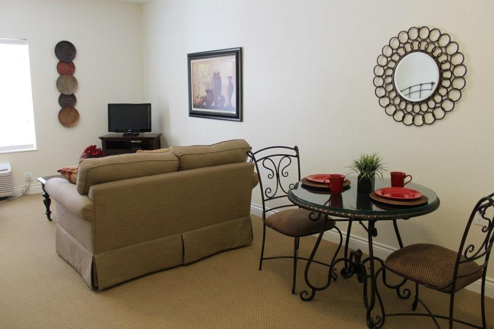 A cozy living room with a small dining area, featuring a sofa, TV, round table with chairs, and decorative items like a mirror and painting.