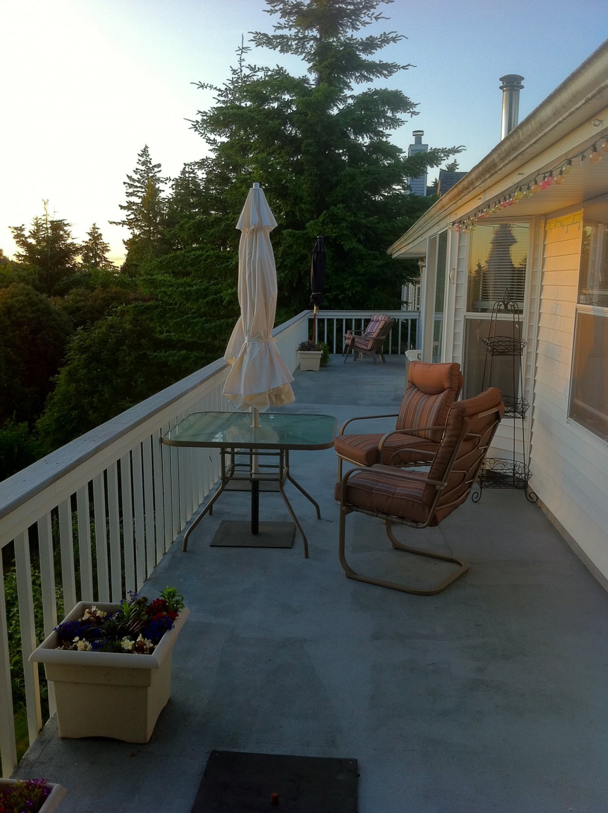 A peaceful balcony setting with chairs, a table, and an umbrella overlooking lush trees at sunset.