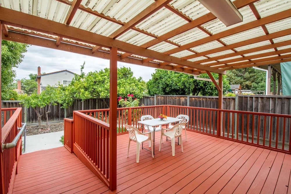 A cozy outdoor patio with a wooden deck and pergola, featuring a set of chairs and a table, surrounded by greenery.