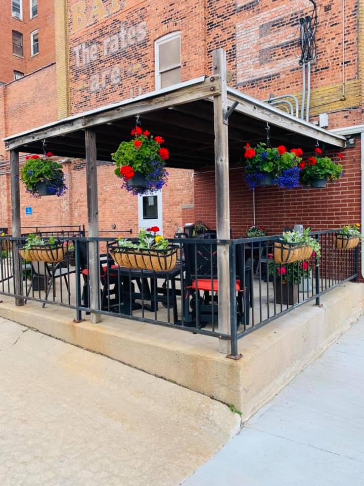 An outdoor patio area with tables and chairs, decorated with hanging plants and located next to a brick building.