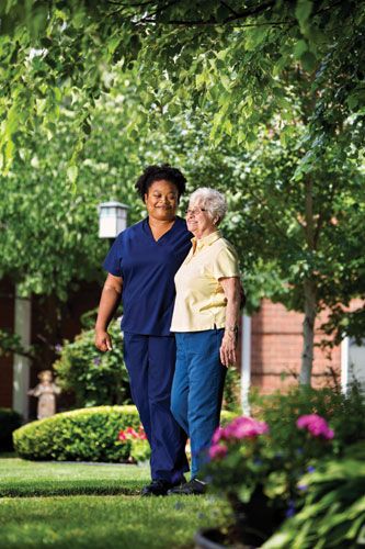 A caregiver walking with an elderly woman in a garden setting.