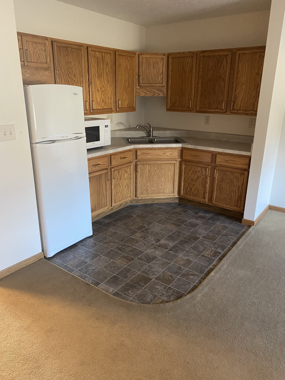 An empty apartment kitchen with wooden cabinets, a white fridge, and a microwave.
