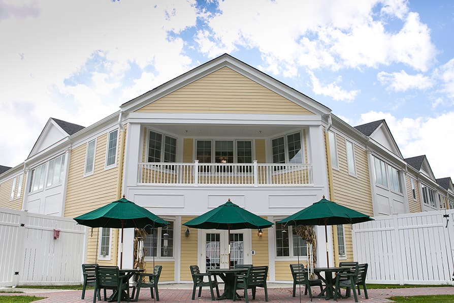 A yellow two-story building with a patio, featuring tables and green umbrellas for outdoor seating under a partly cloudy sky.
