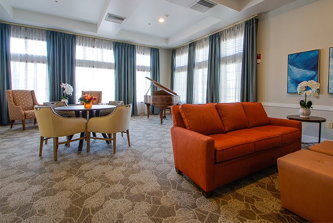 A spacious living room featuring an orange sofa and a grand piano, with large windows adorned with curtains and a patterned carpet.