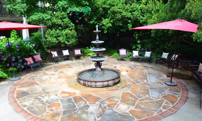 A tranquil outdoor patio with a central multi-tiered fountain surrounded by stone flooring and chairs, shaded by greenery.