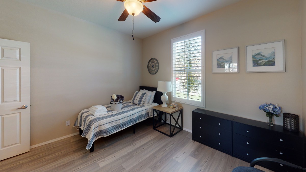 A minimalist bedroom featuring a single bed with striped bedding, a side table with a lamp, and a chest of drawers under sunlight filtering through window shutters.