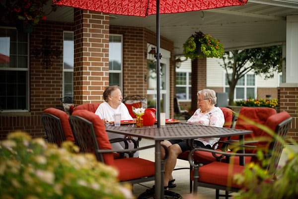 Two elderly women enjoying a conversation under a red umbrella on a patio.