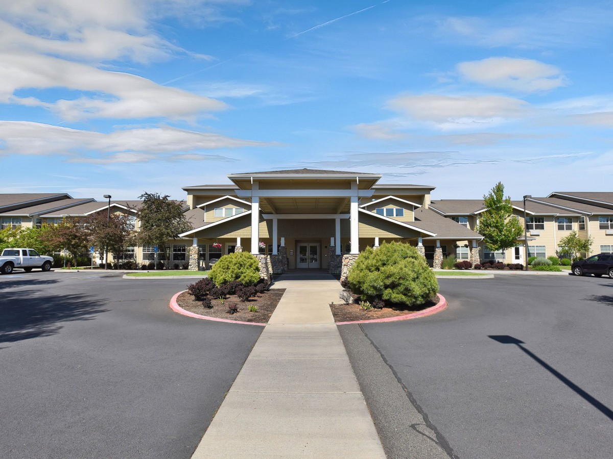 A wide pathway leads to the entrance of a large building, surrounded by neatly trimmed shrubs and landscaping under a partly cloudy sky.