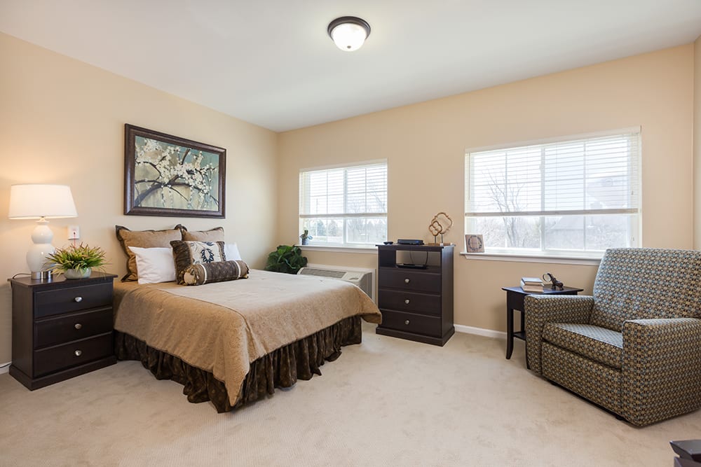 A cozy bedroom with beige walls, featuring a bed, carpet flooring, and two windows allowing natural light.