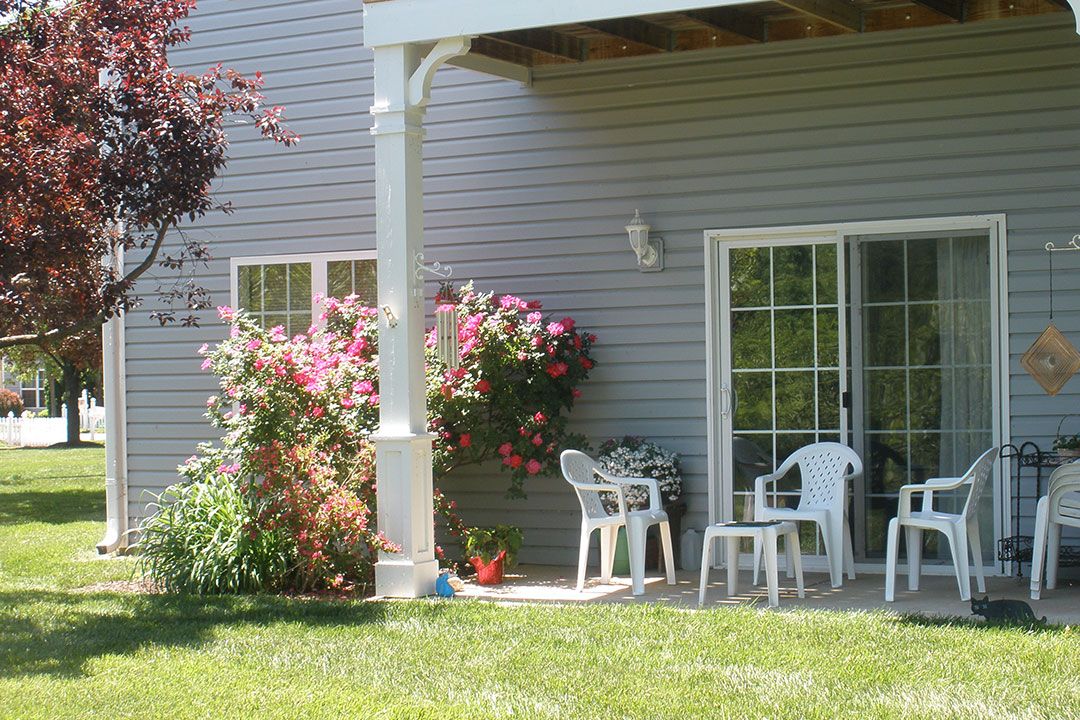A cozy backyard patio with white plastic chairs and a table surrounded by colorful flowers.