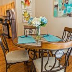 A cozy dining area with a round wooden table, adorned with blue placemats and a floral centerpiece, adjacent to a kitchen space.