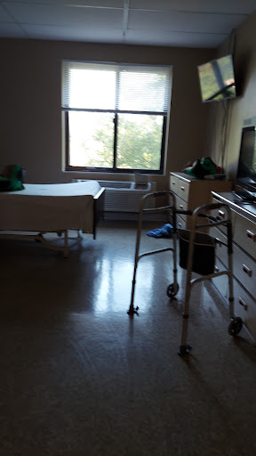 A well-lit hospital room featuring a bed, a walker, and a view outside the window.