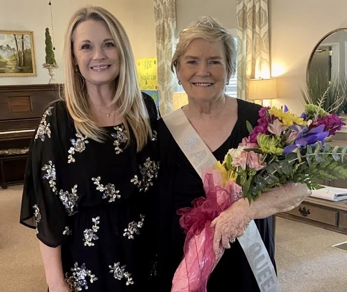 Two women smiling, one holding a bouquet and wearing a sash, during an indoor celebration.