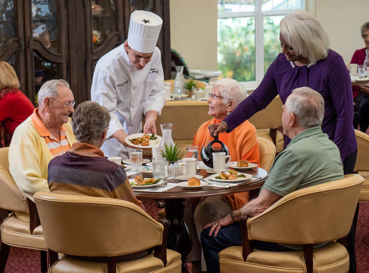 A group of elderly people enjoying a meal together as a chef serves food at their table.