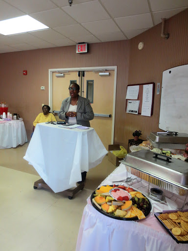 A speaker addresses an audience at a community event, with a buffet table in the foreground.