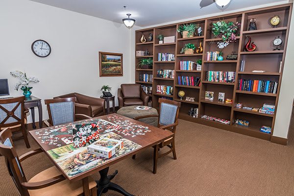 A cozy library room with bookshelves, a table partially covered with a puzzle, and comfortable seating.