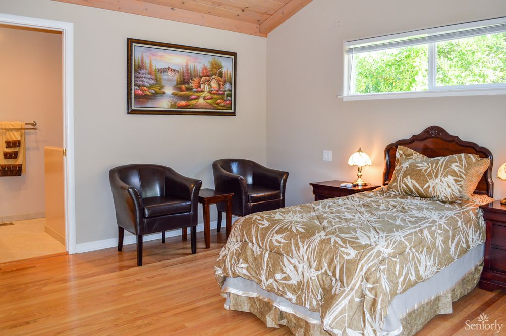 A cozy bedroom with elegant furniture and decor, featuring a bed, two chairs, a lamp, and a painting against a hardwood floor.