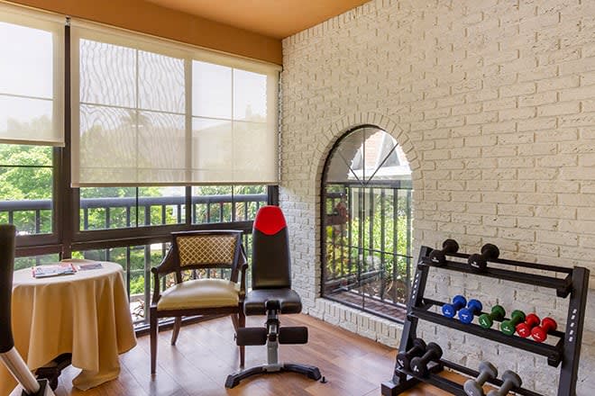 A home gym setup with exercise equipment including weights and a bench, featuring natural light from large windows.