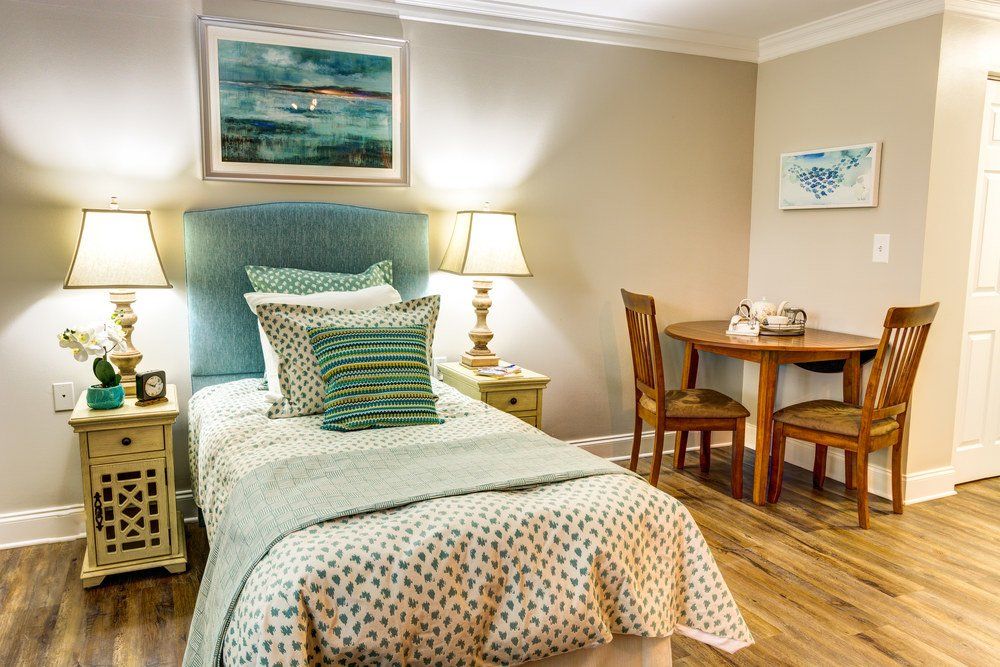 A cozy bedroom with a single bed, decorated with pillows and art, featuring a lamp and small dining table.