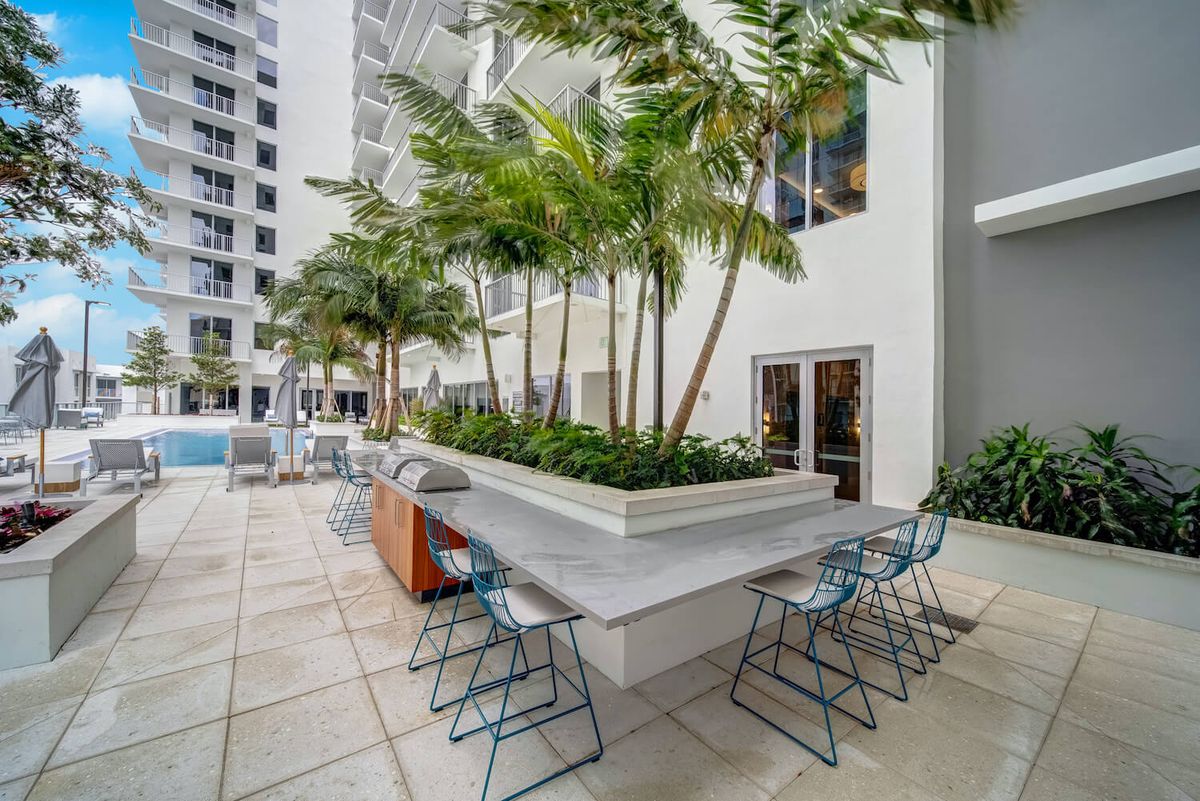 An urban outdoor patio area with tables, chairs, and palm trees surrounded by modern buildings.