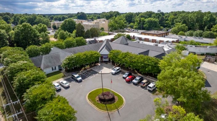 Aerial view of a building with a surrounding parking lot, featuring several cars and lush greenery.