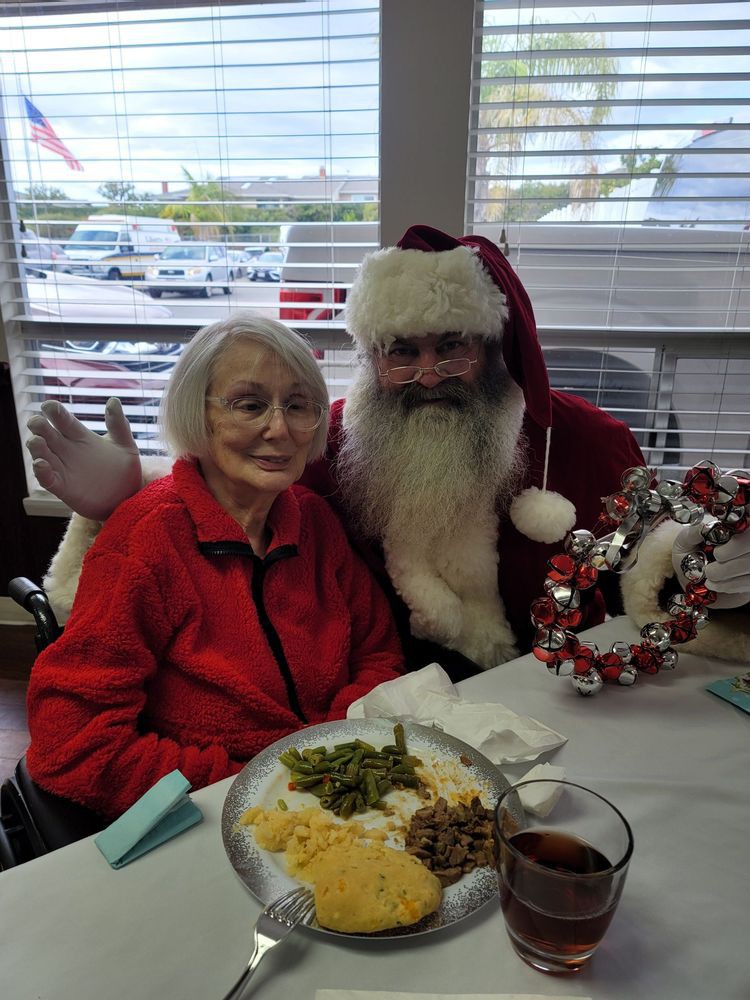 An elderly woman sits with a person dressed as Santa Claus during a festive holiday meal.