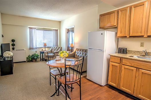 A cozy apartment interior featuring a small kitchen with wooden cabinets, a dining table, and a bright living area.