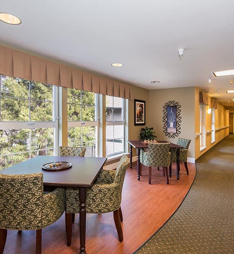 A cozy lounge area with tables and chairs near large windows, adjacent to a hallway.