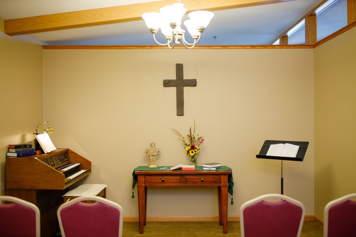 A small chapel room featuring a cross, piano, flowers on a table, and a lectern.