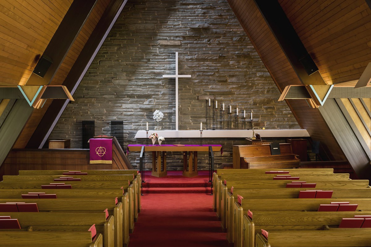 Interior view of a church with wooden ceilings and rows of pews leading to an altar adorned with a cross.