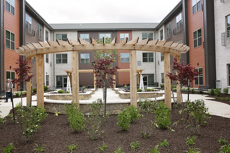 A landscaped courtyard featuring a wooden pergola surrounded by a well-maintained garden and facing a multi-story building.