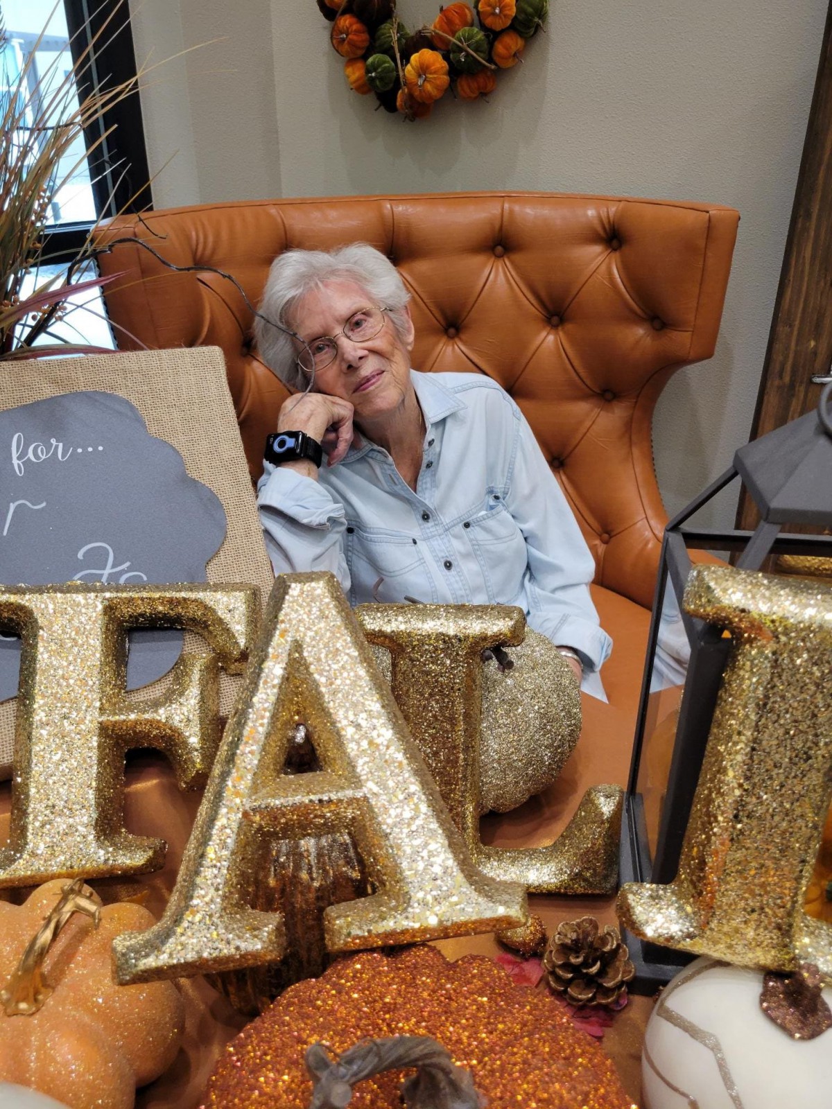 A person smiling and leaning on a chair, surrounded by autumn-themed decorations and large glittery letters spelling 'FALL'.